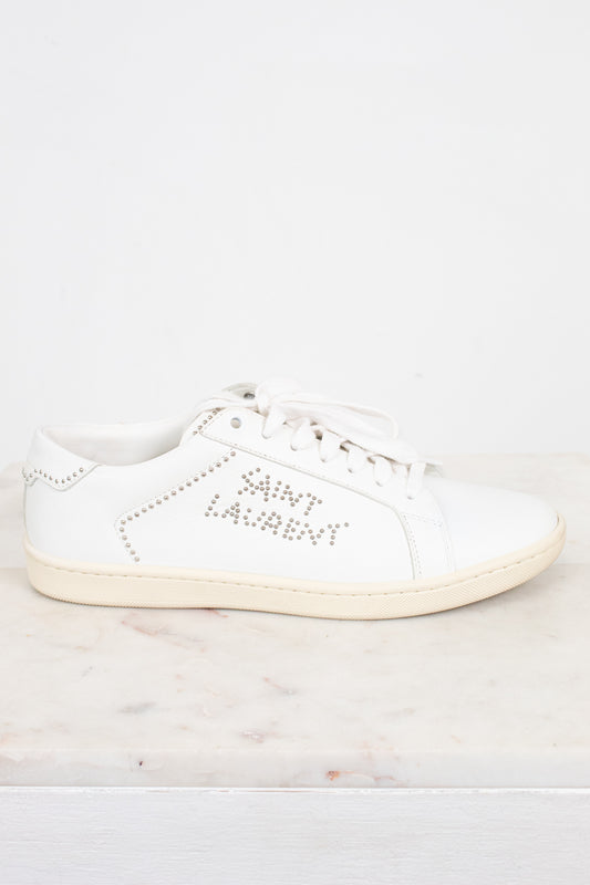 A white leather Saint Laurent sneaker with silver rhinestone lettering on the side, displayed on a light marble surface against a white background.