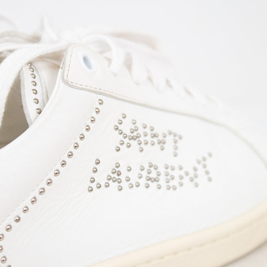 A close-up of a white leather sneaker featuring silver rhinestone lettering that spells 'SAINT LAURENT' and decorative rhinestone borders along the side, with a cream-colored sole.