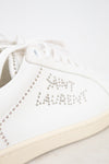 A close-up of a white leather sneaker featuring silver rhinestone lettering that spells 'SAINT LAURENT' and decorative rhinestone borders along the side, with a cream-colored sole.