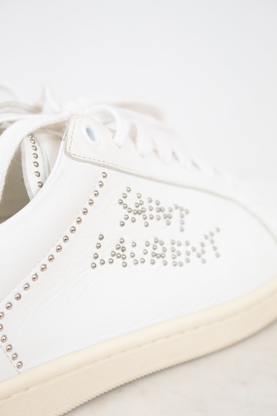A close-up of a white leather sneaker featuring silver rhinestone lettering that spells 'SAINT LAURENT' and decorative rhinestone borders along the side, with a cream-colored sole.