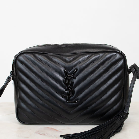 A black quilted leather shoulder bag with a chevron pattern, a black YSL logo on the front, and a black tassel detail, displayed on a light marble surface against a white background.