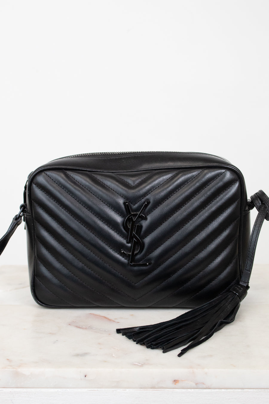 A black quilted leather shoulder bag with a chevron pattern, a black YSL logo on the front, and a black tassel detail, displayed on a light marble surface against a white background.