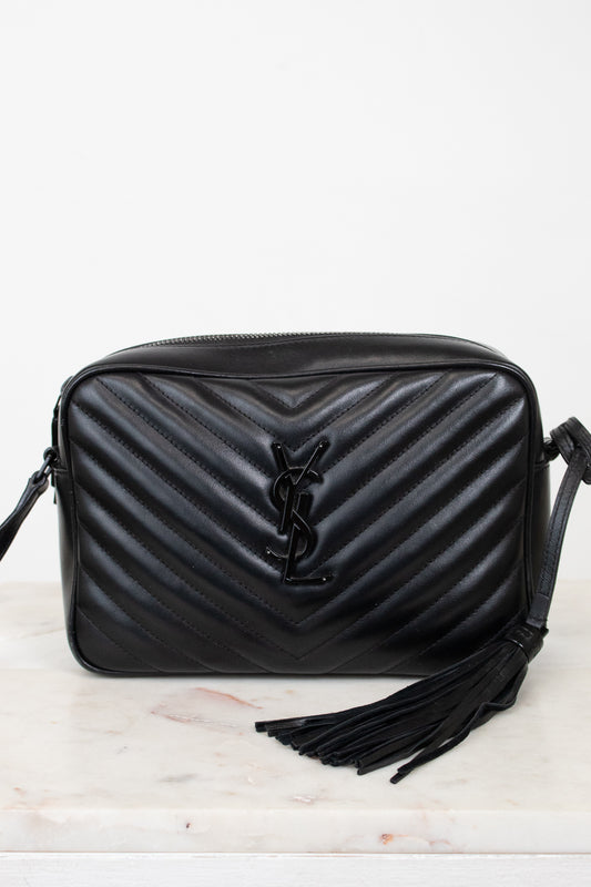 A black quilted leather shoulder bag with a chevron pattern, a black YSL logo on the front, and a black tassel detail, displayed on a light marble surface against a white background.