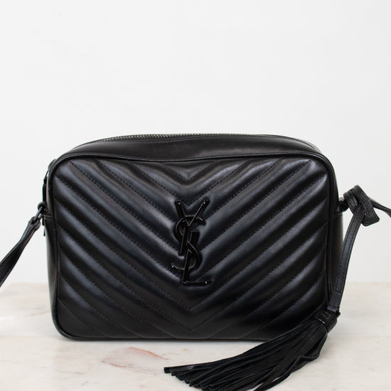 A black quilted leather shoulder bag with a prominent interlocking YSL logo on the front, resting on a light marble surface against a white background.
