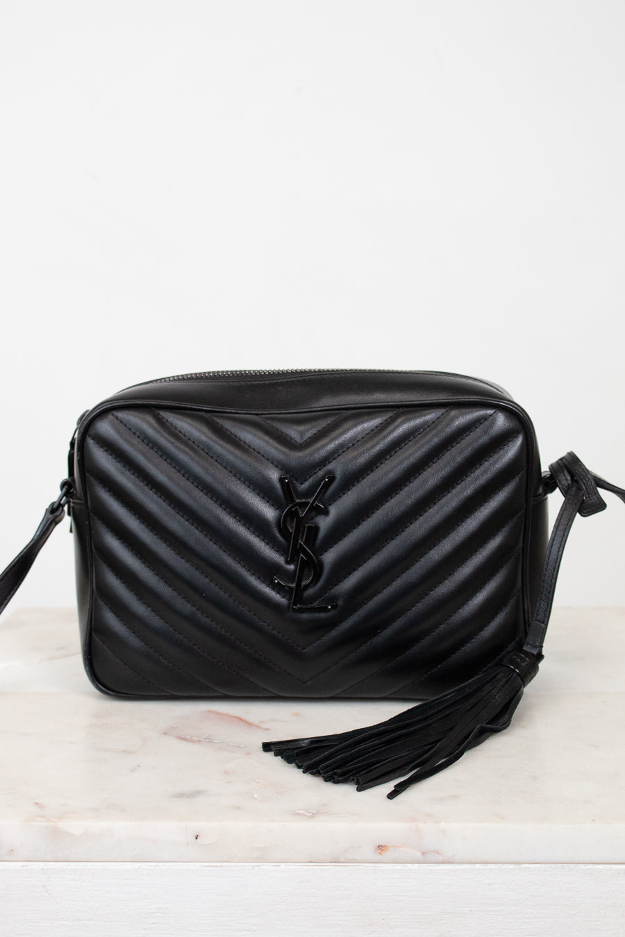 A black quilted leather shoulder bag with a prominent interlocking YSL logo on the front, resting on a light marble surface against a white background.