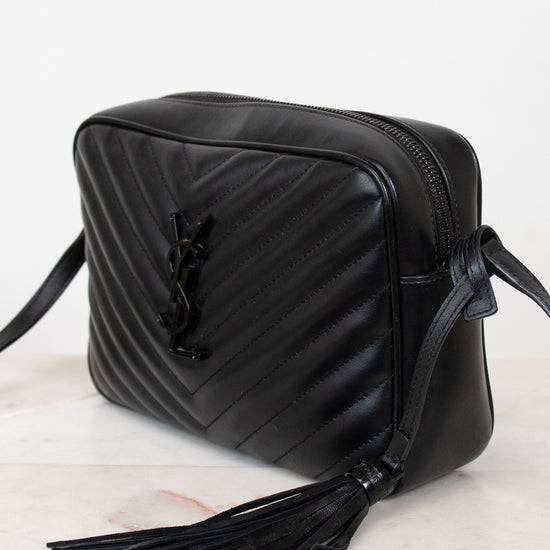 A black leather crossbody bag with a diagonal chevron quilted pattern, a black leather tassel, and a black metal YSL logo clasp, resting on a light marble surface against a white background.