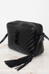 A black leather crossbody bag with a diagonal chevron quilted pattern, a black leather tassel, and a black metal YSL logo clasp, resting on a light marble surface against a white background.