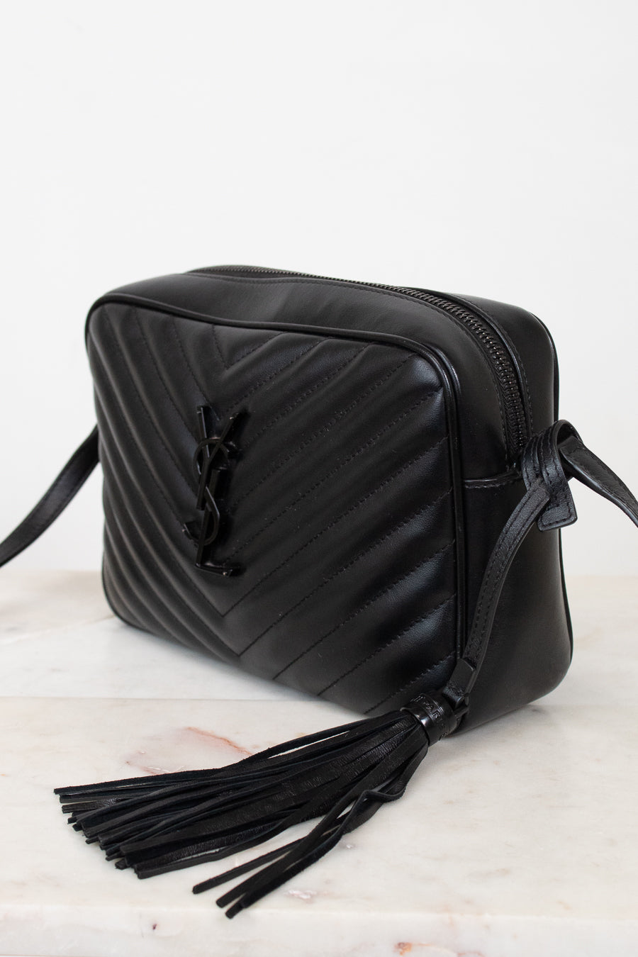 A black leather crossbody bag with a diagonal chevron quilted pattern, a black leather tassel, and a black metal YSL logo clasp, resting on a light marble surface against a white background.