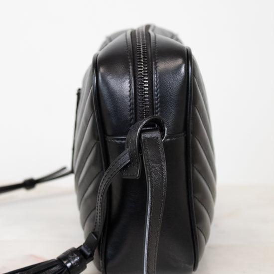 A black quilted leather crossbody bag with a tassel detail, shown from a side profile view on a light marble surface against a white background.