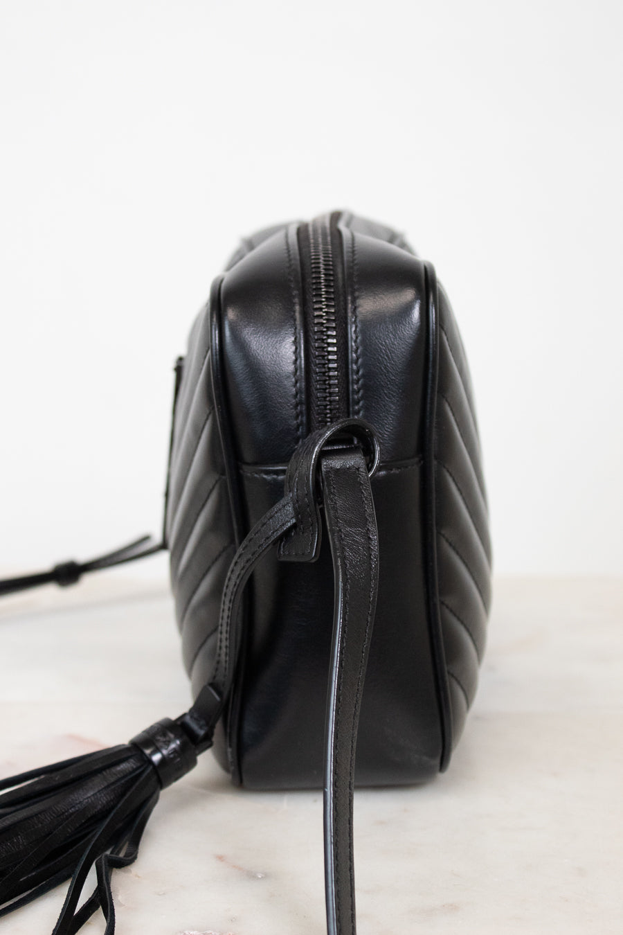 A black quilted leather crossbody bag with a tassel detail, shown from a side profile view on a light marble surface against a white background.