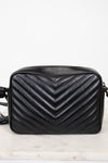 A black quilted leather crossbody bag with a prominent chevron pattern, resting on a light marble surface against a white background.