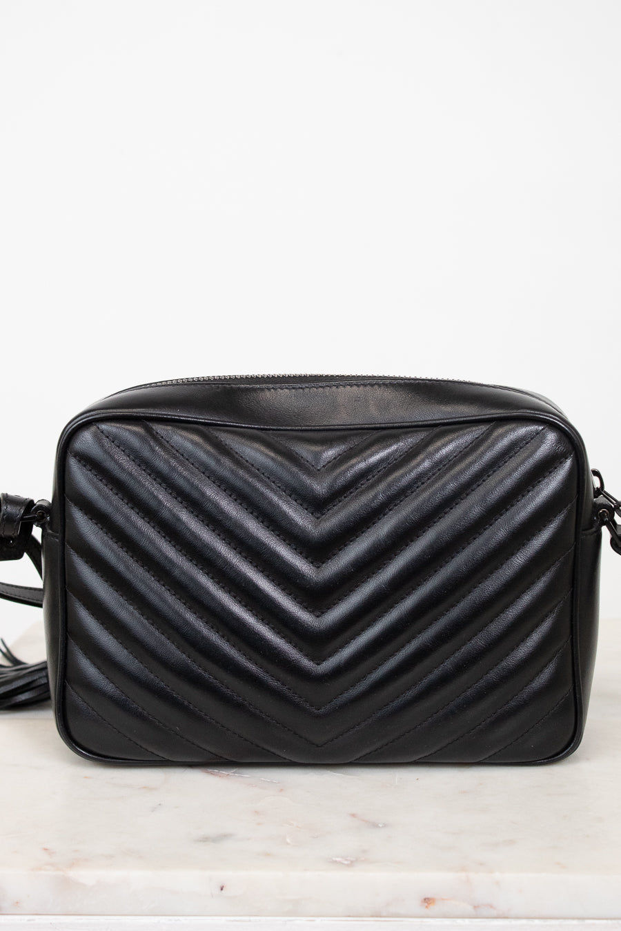 A black quilted leather crossbody bag with a prominent chevron pattern, resting on a light marble surface against a white background.