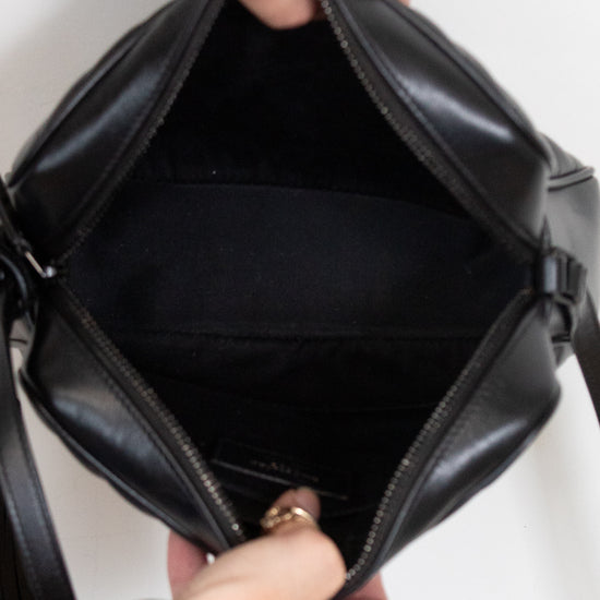 A close-up of a black leather handbag being opened by hands, showing the interior lining and zipper. The person's hands, adorned with rings and bracelets, are visible against a plain white background.