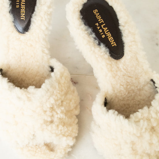 A pair of off-white slide slippers with black insoles featuring gold Saint Laurent Paris branding, resting on a light marble surface.