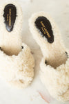 A pair of off-white slide slippers with black insoles featuring gold Saint Laurent Paris branding, resting on a light marble surface.