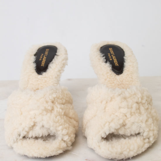 A pair of cream-colored, fluffy shearling-style slide sandals with black insoles, displayed on a light marble surface against a white background.