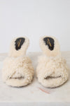 A pair of cream-colored, fluffy shearling-style slide sandals with black insoles, displayed on a light marble surface against a white background.