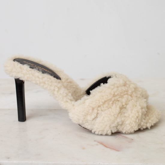 A single cream-colored shearling mule sandal with a black stiletto heel, resting on a white marble surface against a white background.