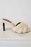 A single cream-colored shearling mule sandal with a black stiletto heel, resting on a white marble surface against a white background.