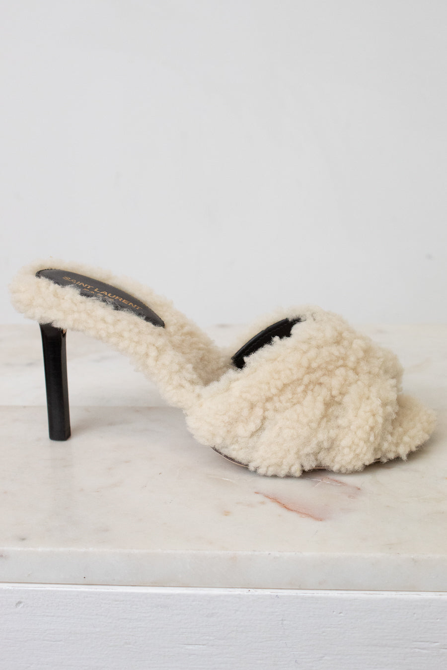 A single cream-colored shearling mule sandal with a black stiletto heel, resting on a white marble surface against a white background.