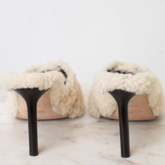 A pair of off-white shearling slip-on mules with black stiletto heels, shown from the back on a light marble surface against a white background.