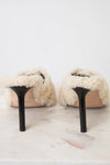 A pair of off-white shearling slip-on mules with black stiletto heels, shown from the back on a light marble surface against a white background.