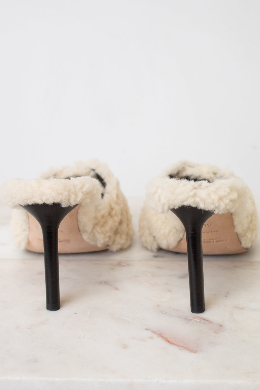 A pair of off-white shearling slip-on mules with black stiletto heels, shown from the back on a light marble surface against a white background.