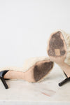 A pair of black high-heeled shoes with thick cream-colored shearling uppers, photographed from below on a light marble surface against a white background.