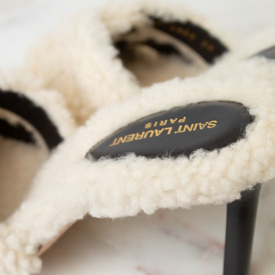 A close-up of a Saint Laurent high-heeled slide sandal featuring a cream-colored shearling upper and a black leather insole with gold lettering, resting on a light marble surface.