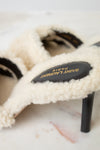 A close-up of a Saint Laurent high-heeled slide sandal featuring a cream-colored shearling upper and a black leather insole with gold lettering, resting on a light marble surface.