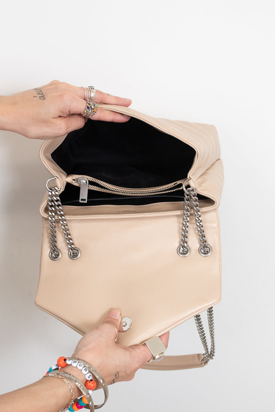 A beige leather shoulder bag with silver chain details and a black interior, held open by two hands against a plain white background.