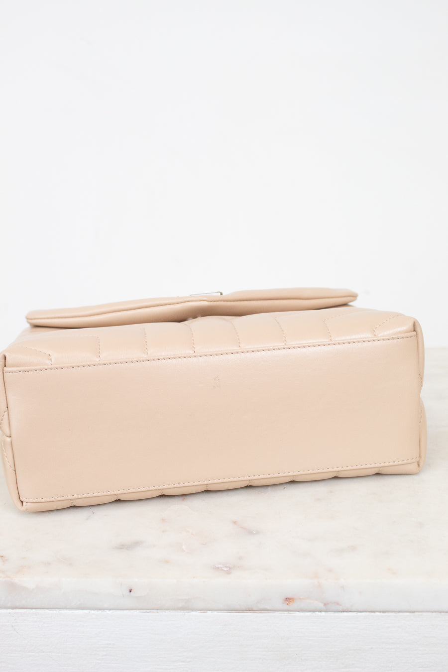 A light beige leather bag with a quilted top flap, resting on a white marble surface against a white background.