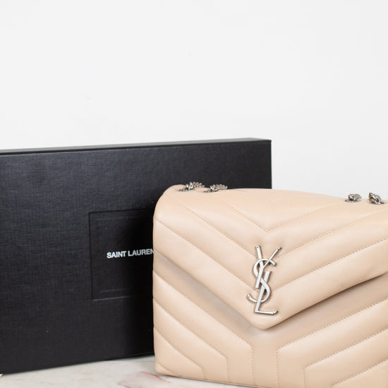 A Saint Laurent shoulder bag in beige pebbled leather with a silver-tone YSL logo clasp, shown next to its black Saint Laurent box on a light marble surface.