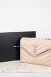 A Saint Laurent shoulder bag in beige pebbled leather with a silver-tone YSL logo clasp, shown next to its black Saint Laurent box on a light marble surface.