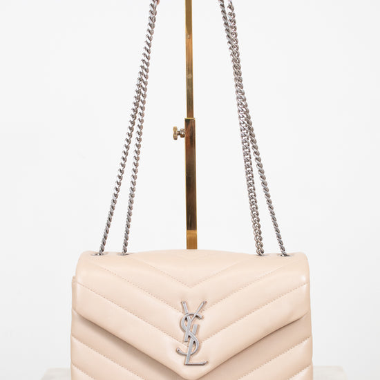 A light beige quilted leather shoulder bag with a silver-tone YSL monogram clasp, displayed on a gold stand with a white background.