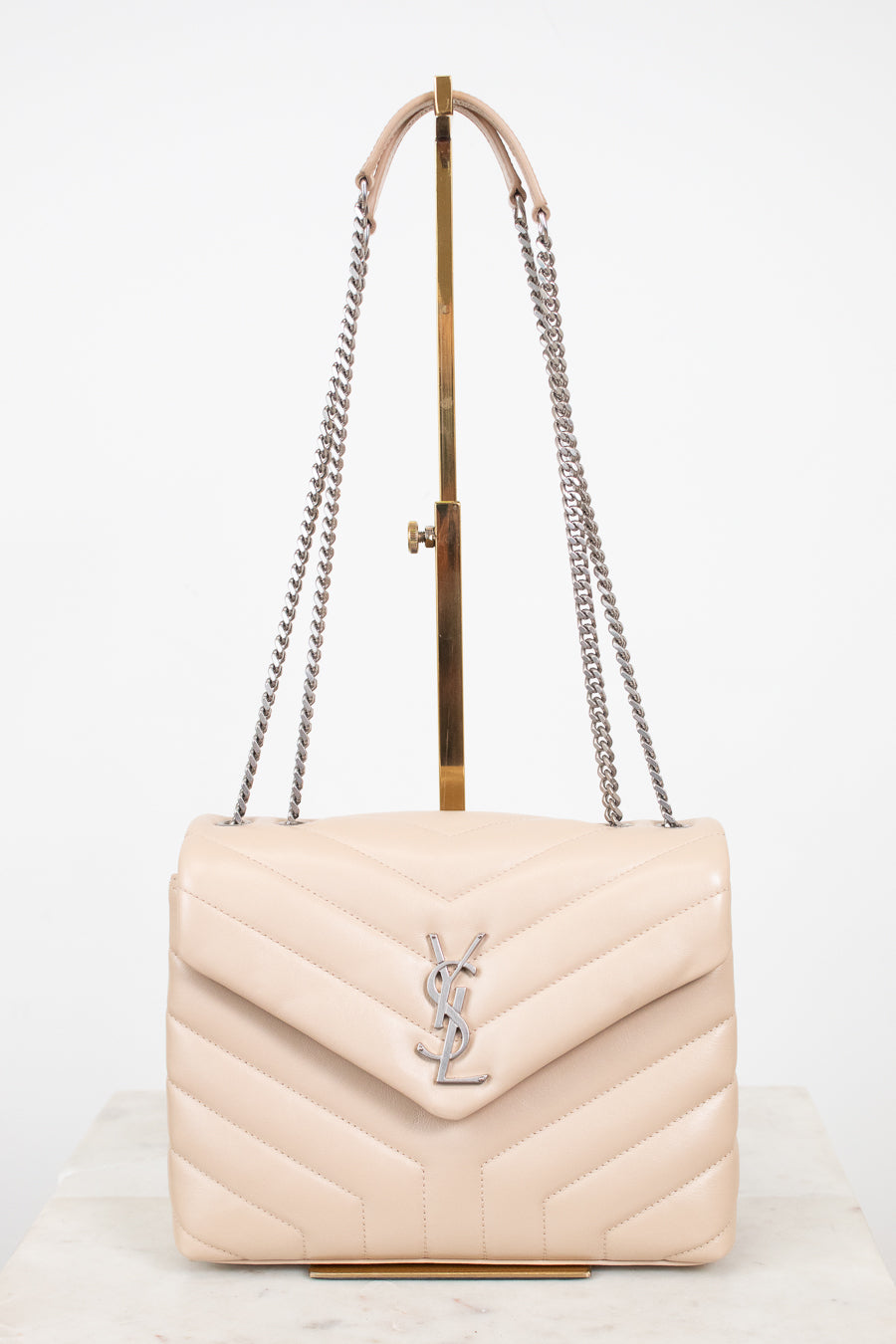 A light beige quilted leather shoulder bag with a silver-tone YSL monogram clasp, displayed on a gold stand with a white background.