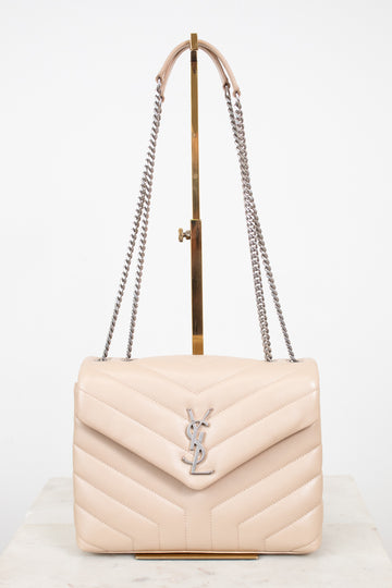 A light beige quilted leather shoulder bag with a silver-tone YSL monogram clasp, displayed on a gold stand with a white background.