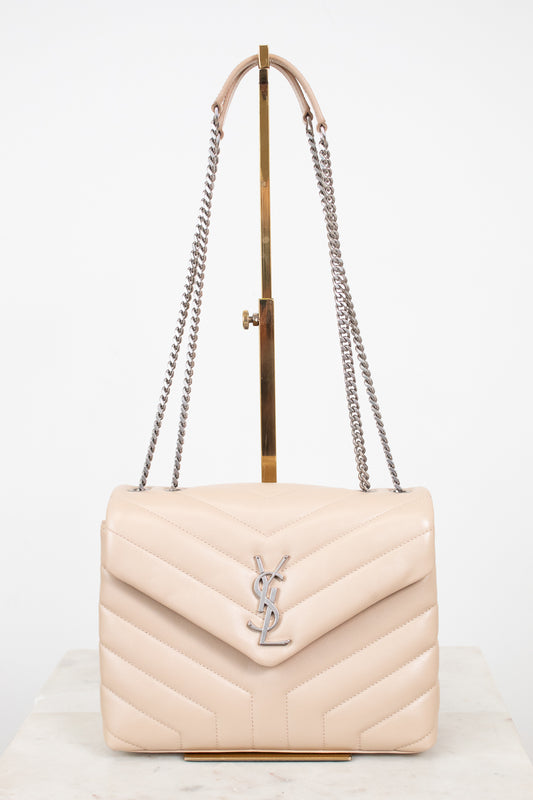 A light beige quilted leather shoulder bag with a silver-tone YSL monogram clasp, displayed on a gold stand with a white background.