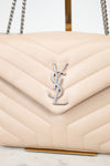 A close-up of a quilted cream-colored leather handbag with a silver-tone YSL logo on the front flap and silver chain straps, resting on a marble surface.