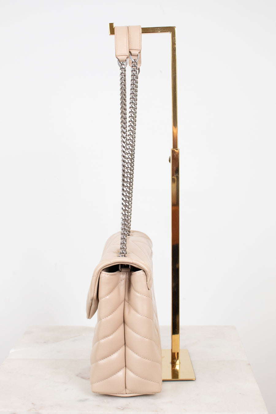 A light beige quilted leather shoulder bag with a silver chain strap, displayed on a gold metal stand against a white background.