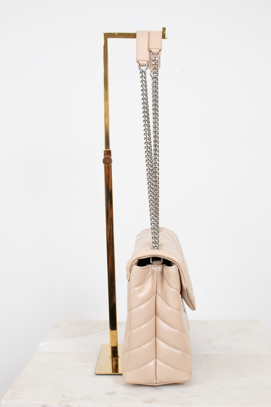 A light beige quilted leather shoulder bag with a silver chain strap, displayed on a gold metal stand against a white background.