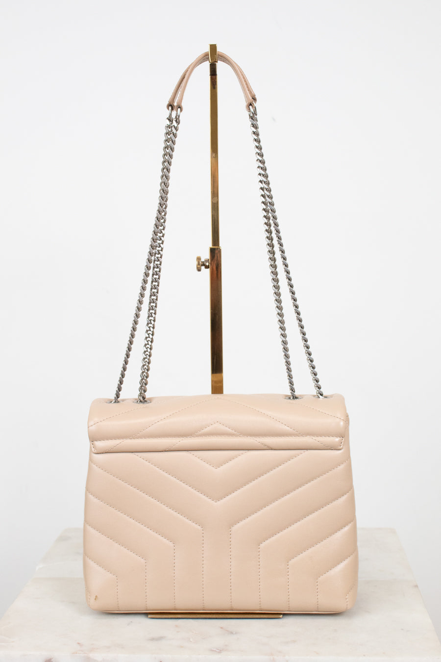 A light beige quilted leather shoulder bag with a geometric chevron pattern, featuring silver-tone chain straps and a gold-tone metal handle, displayed on a stand.