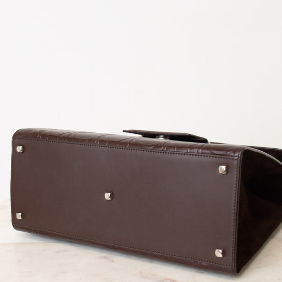 A dark brown leather handbag shown from a low angle, highlighting its bottom corner with silver-tone studs, resting on a light marble surface against a white background.