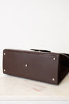 A dark brown leather handbag shown from a low angle, highlighting its bottom corner with silver-tone studs, resting on a light marble surface against a white background.