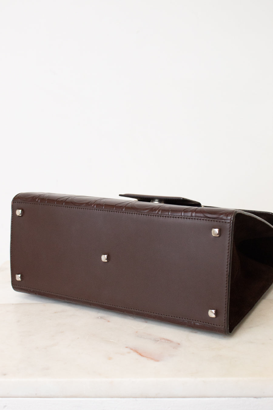 A dark brown leather handbag shown from a low angle, highlighting its bottom corner with silver-tone studs, resting on a light marble surface against a white background.