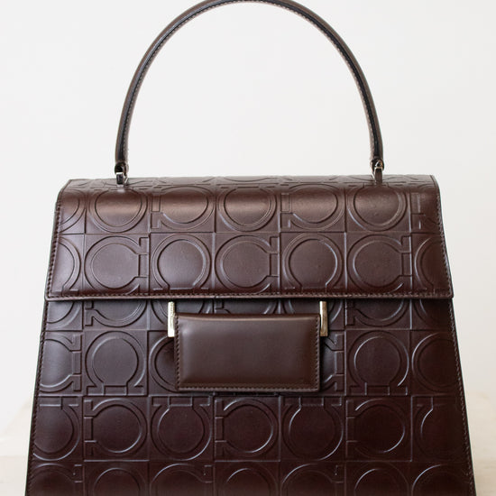 A dark brown leather handbag with an embossed geometric circle pattern, featuring a top handle, a front flap pocket, and small gold-tone hardware, shown against a plain white background.
