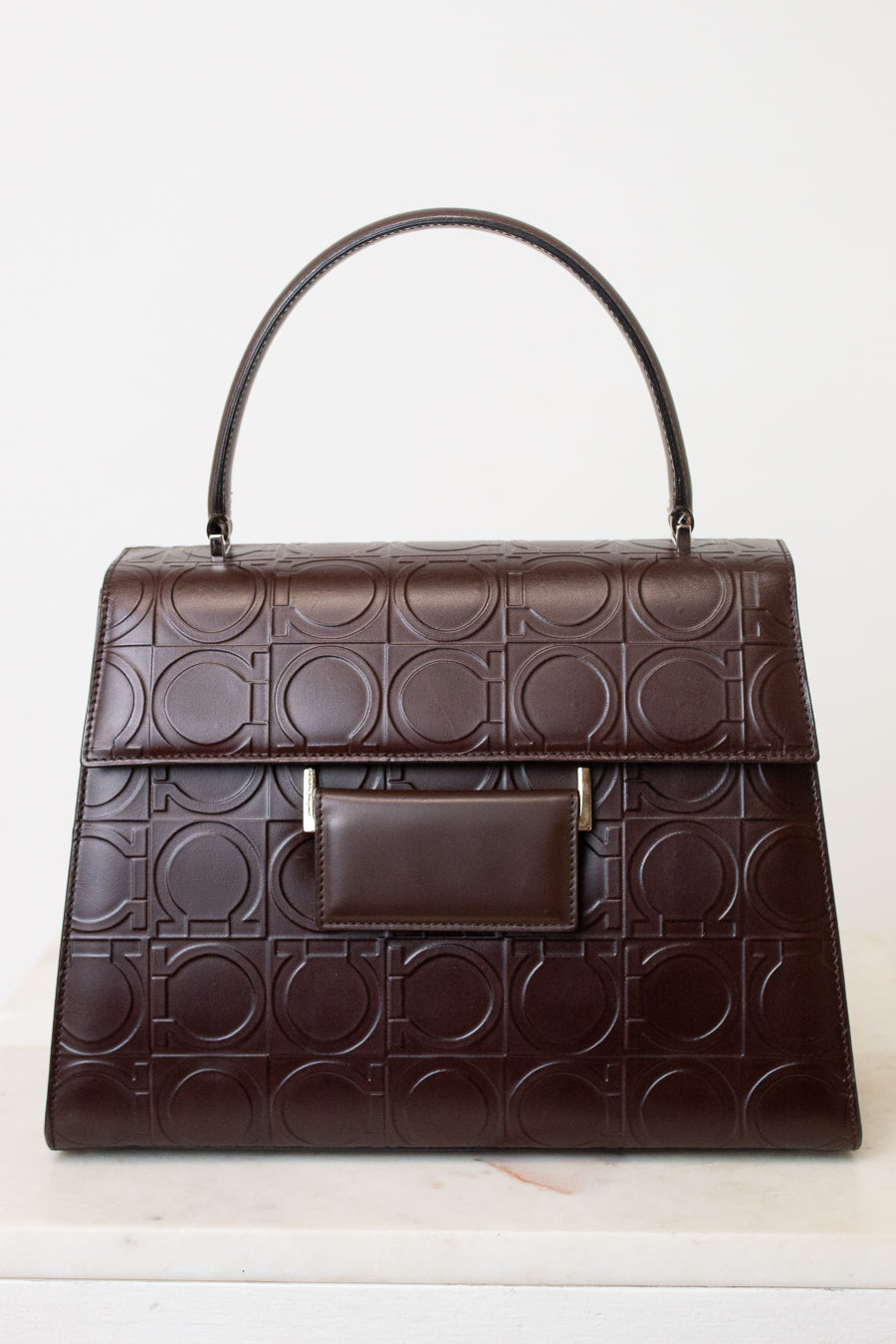 A dark brown leather handbag with an embossed geometric circle pattern, featuring a top handle, a front flap pocket, and small gold-tone hardware, shown against a plain white background.