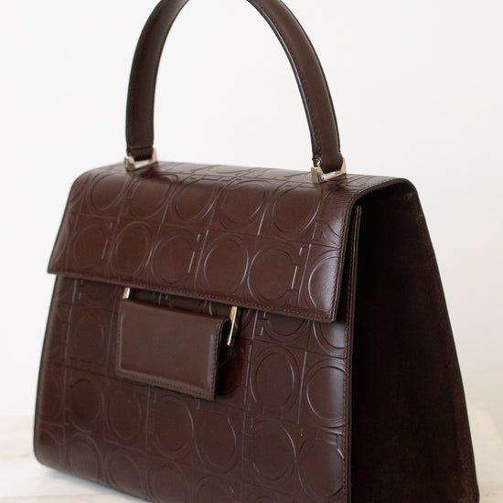A dark brown leather handbag with an embossed circular pattern, featuring a top handle, a front flap pocket, and gold-tone hardware, set against a plain white background.