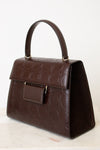 A dark brown leather handbag with an embossed circular pattern, featuring a top handle, a front flap pocket, and gold-tone hardware, set against a plain white background.
