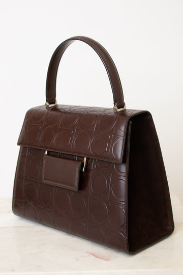 A dark brown leather handbag with an embossed circular pattern, featuring a top handle, a front flap pocket, and gold-tone hardware, set against a plain white background.